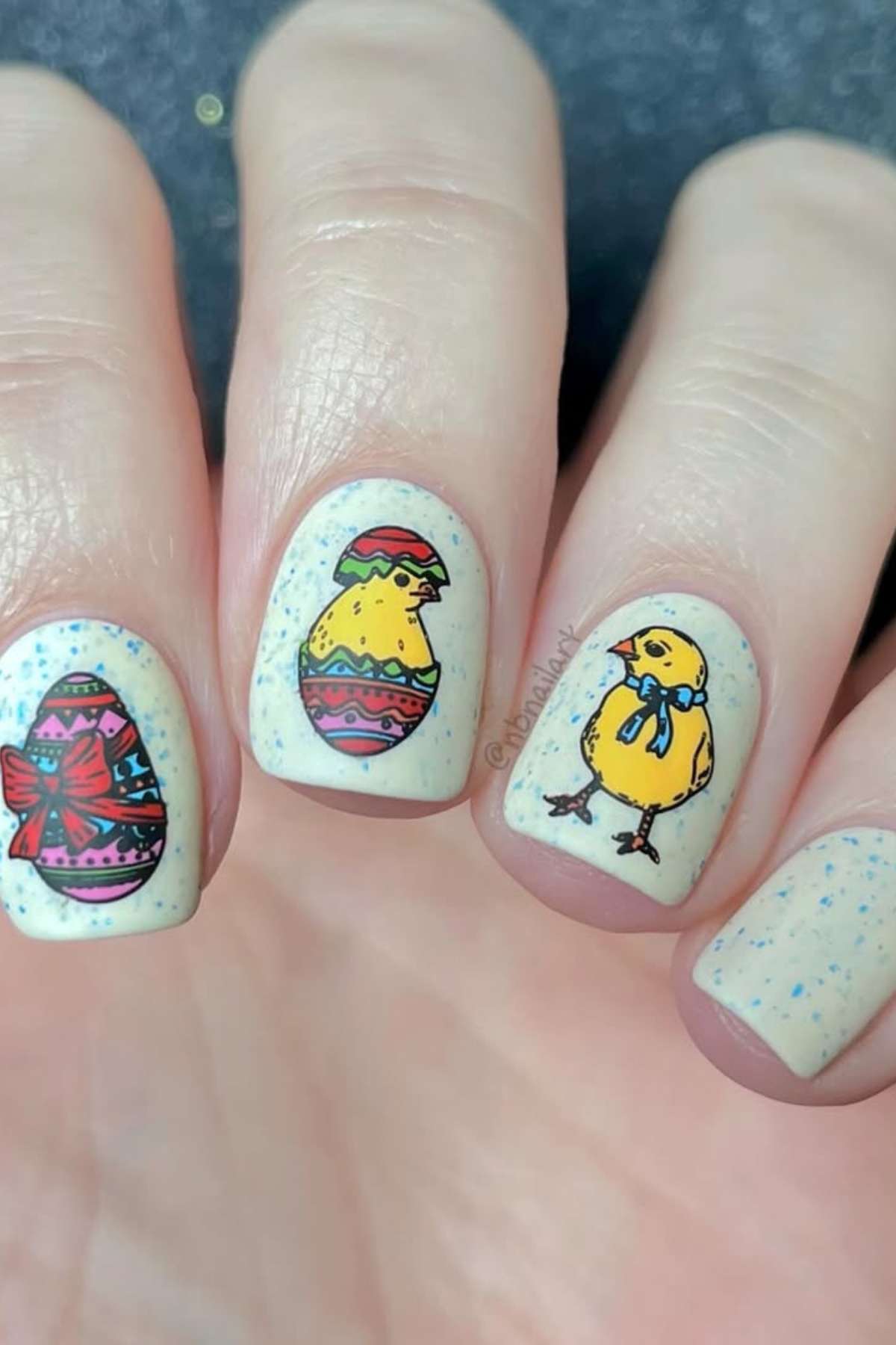 A close-up of fingernails painted with Easter-themed designs, including decorated eggs and yellow chicks, on a speckled light background.