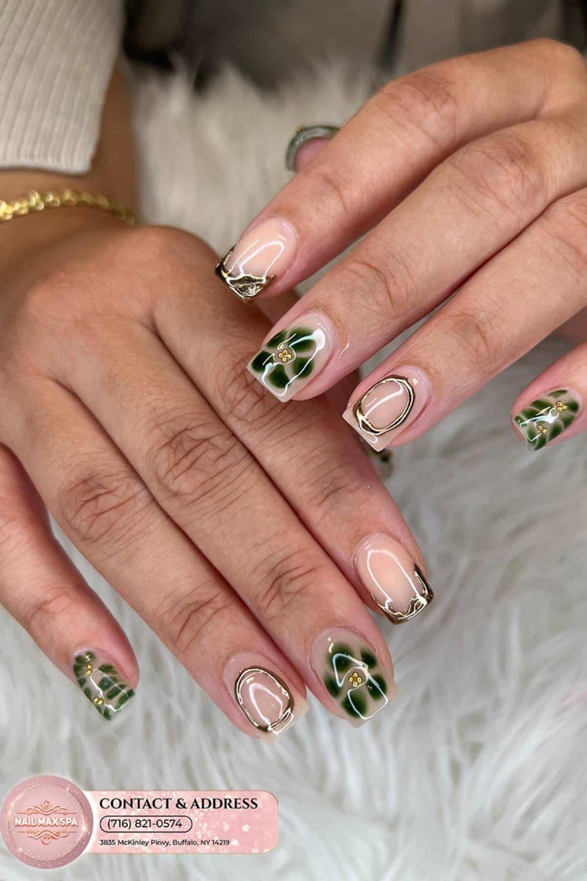 A close-up of manicured hands with green and gold floral nail art and gold accents, resting on a white textured surface.