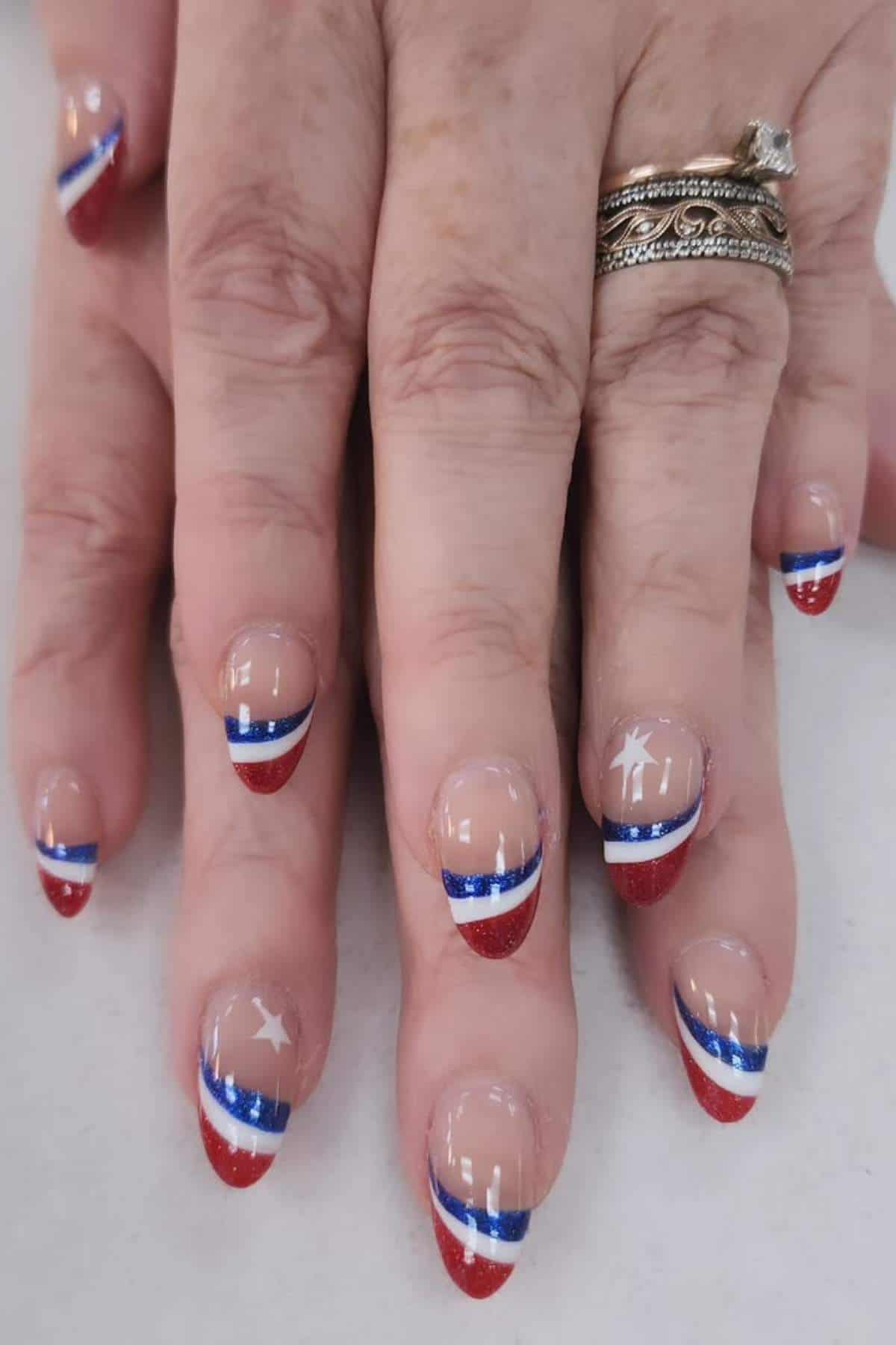 Close-up of hands with manicured nails decorated in red, white, and blue stripes, some featuring white stars. The person is wearing multiple rings.