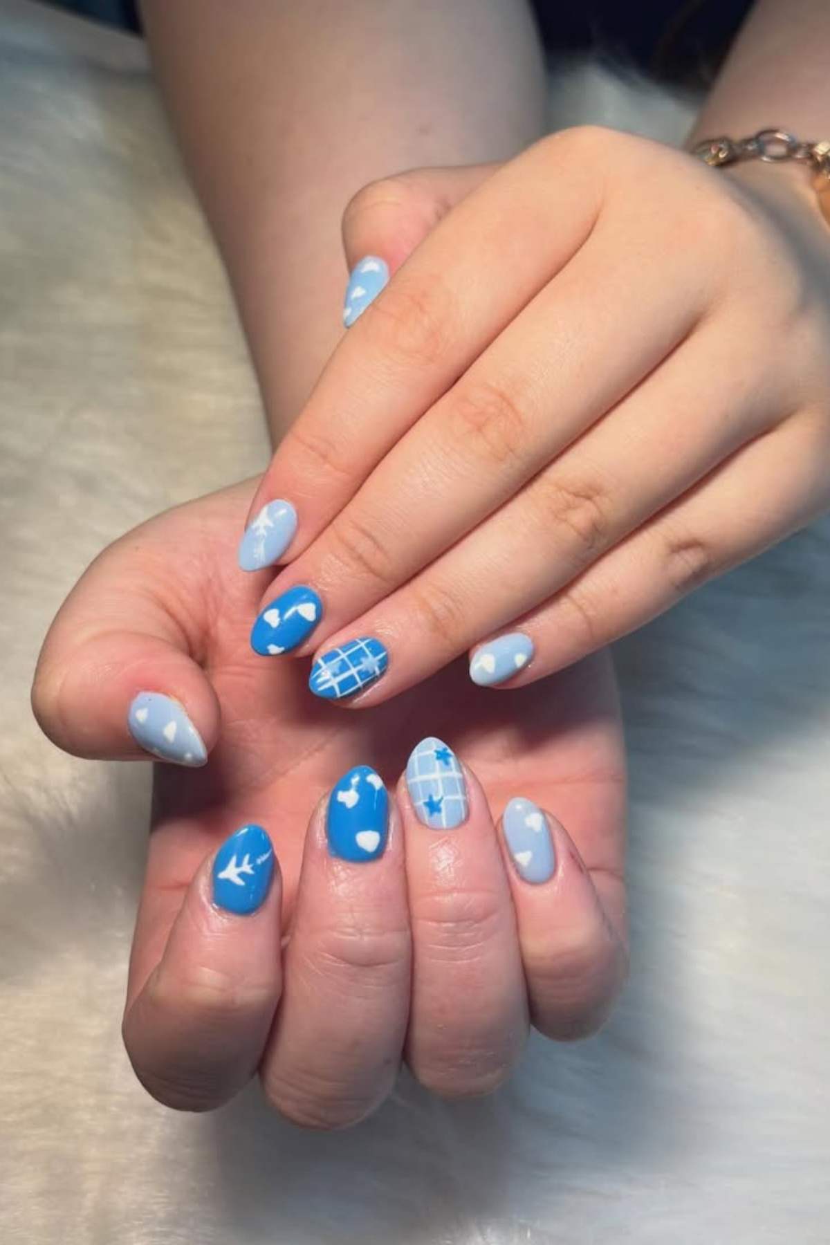 Hands with manicured nails in blue and light blue shades, featuring designs of hearts, clouds, a grid pattern, and a bird on a soft white surface.