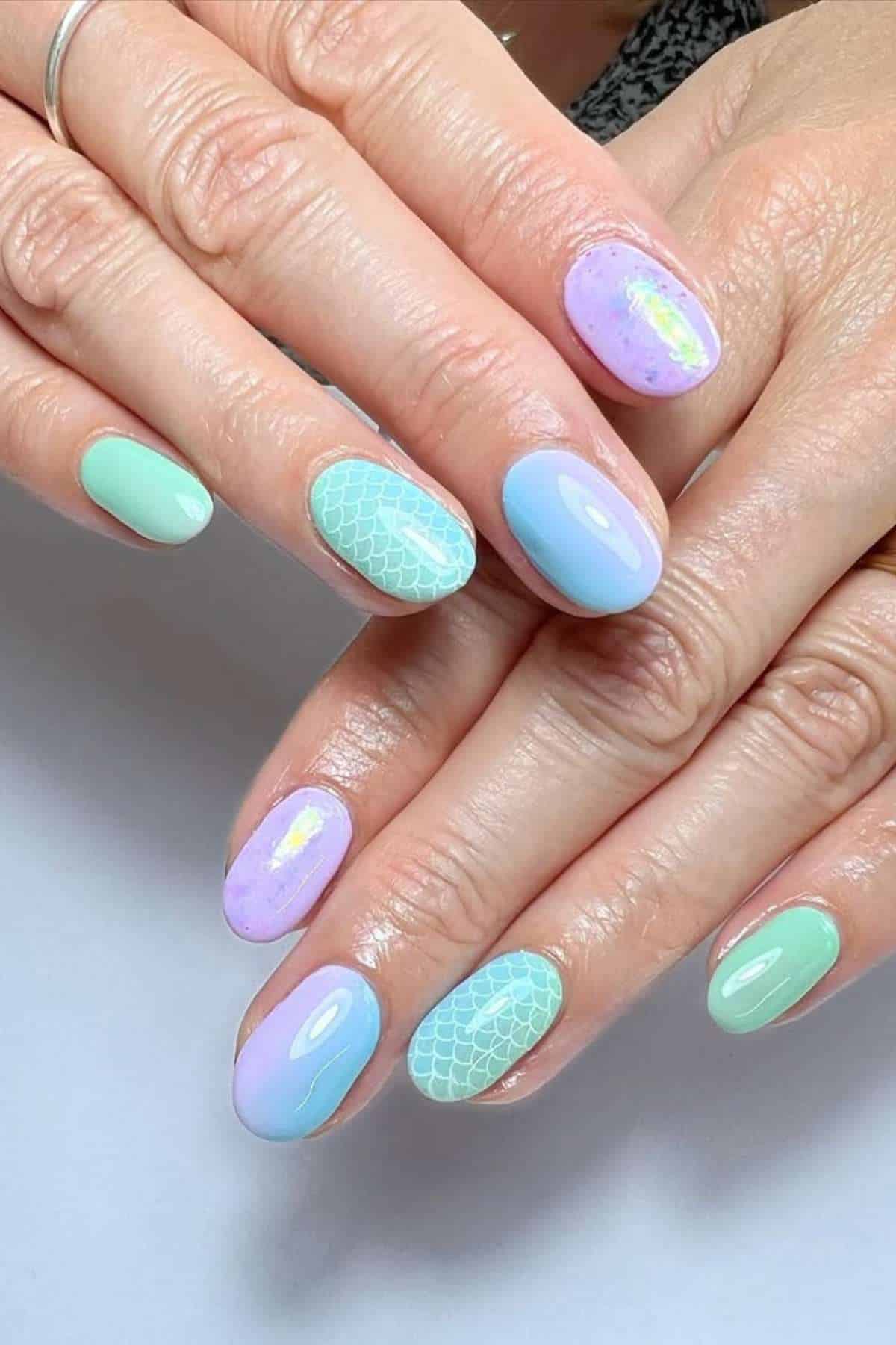 Hands with pastel-colored manicure featuring light green, blue, and purple shades; some nails have mermaid scale patterns and others have iridescent accents.