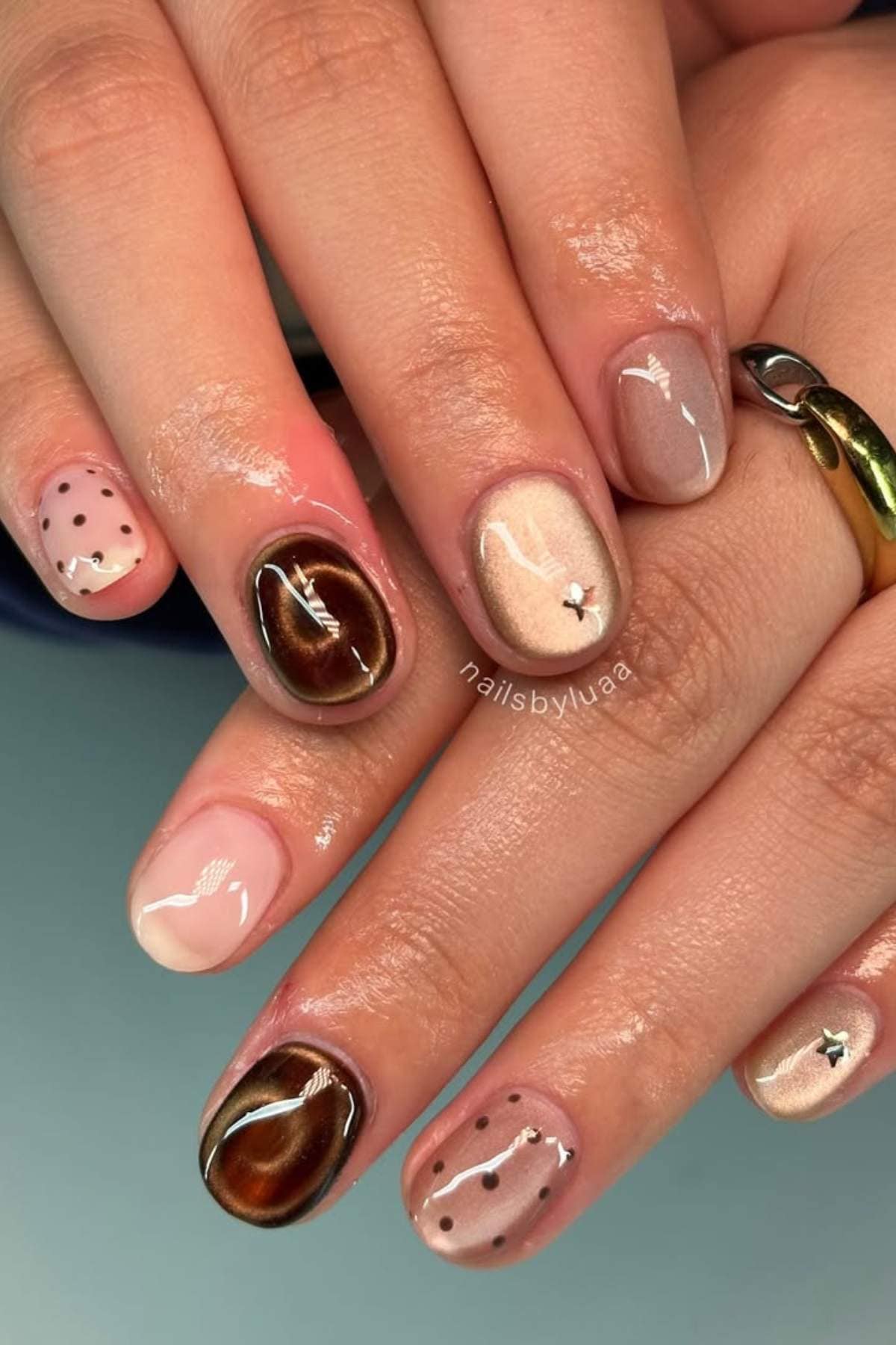 A hand with almond-shaped nails featuring a mix of brown marble, nude, and taupe polishes, some with polka dots and small metallic star accents.