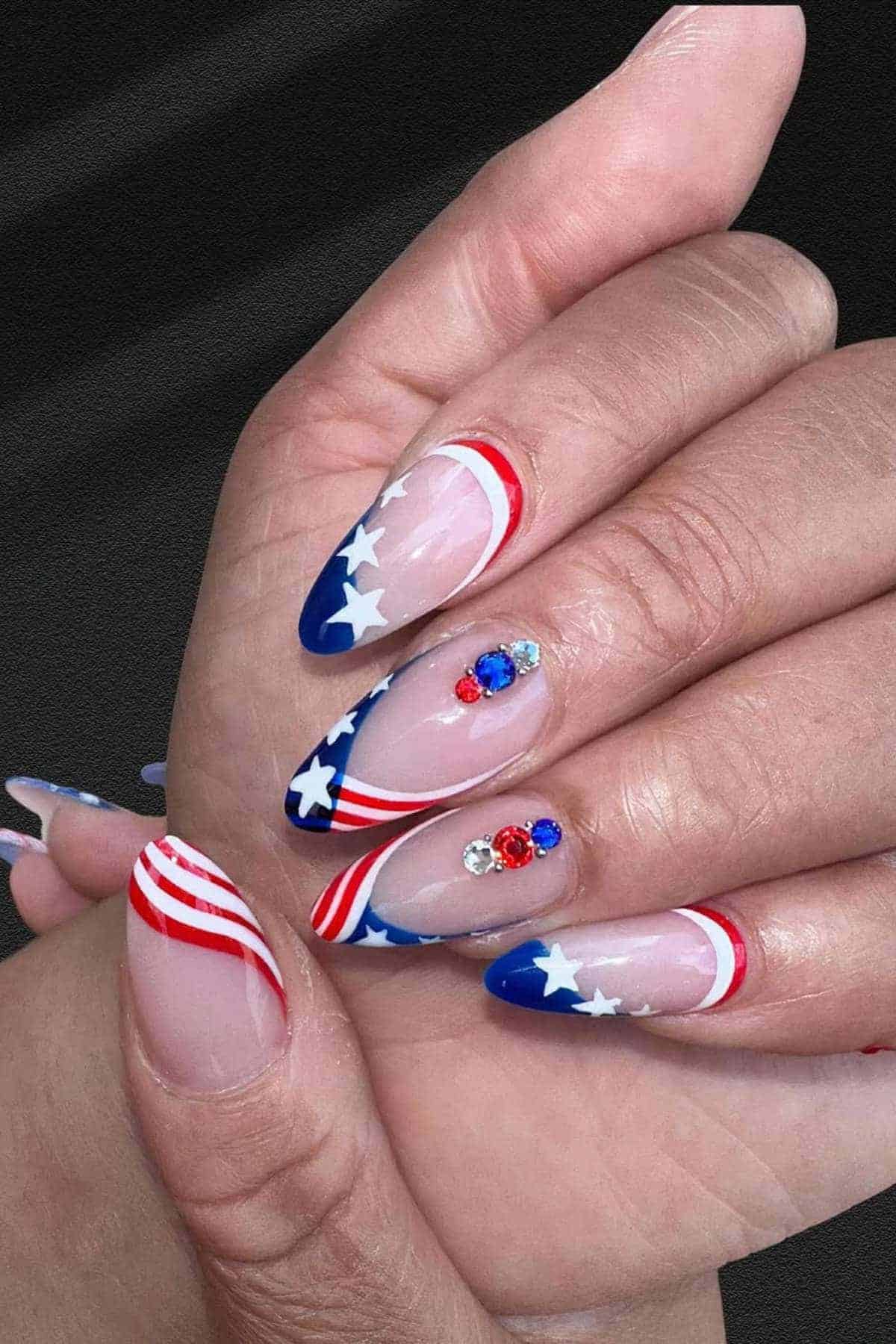 Hands with almond-shaped nails featuring an American flag design with red, white, and blue patterns, stars, and rhinestone accents.