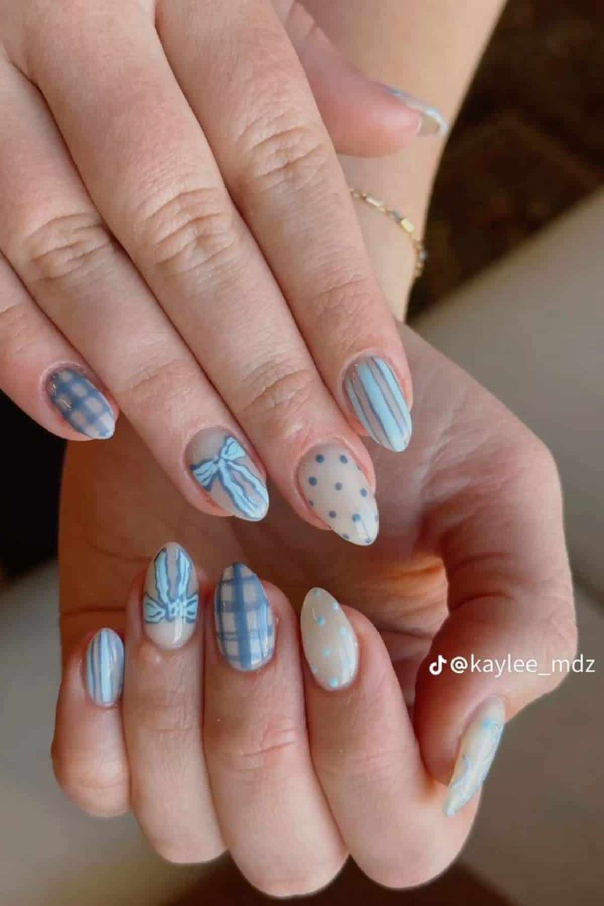 Hands display a pastel blue manicure with various designs including stripes, plaid, polka dots, and bows on oval-shaped nails.