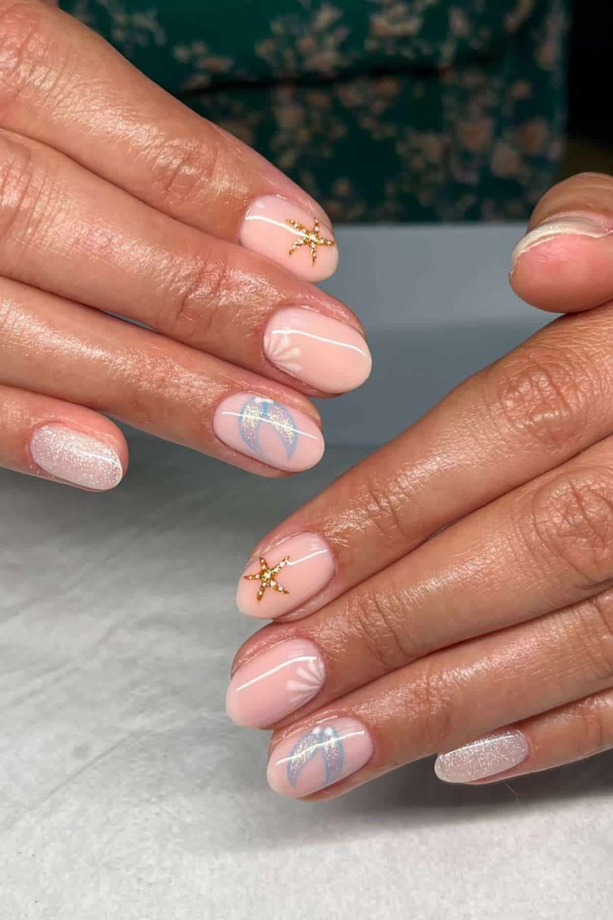 Hands with short, oval-shaped nails painted light pink, featuring gold star and silver leaf nail art on some fingers, resting on a white surface.