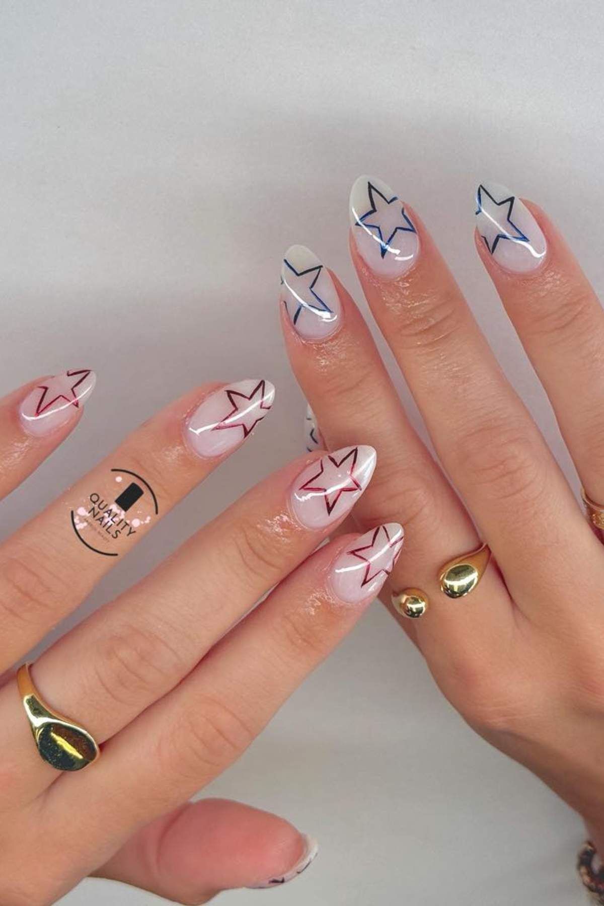 Hands with almond-shaped nails featuring minimalist star designs in black, red, and blue; wearer has three gold rings and a small temporary tattoo on one finger.