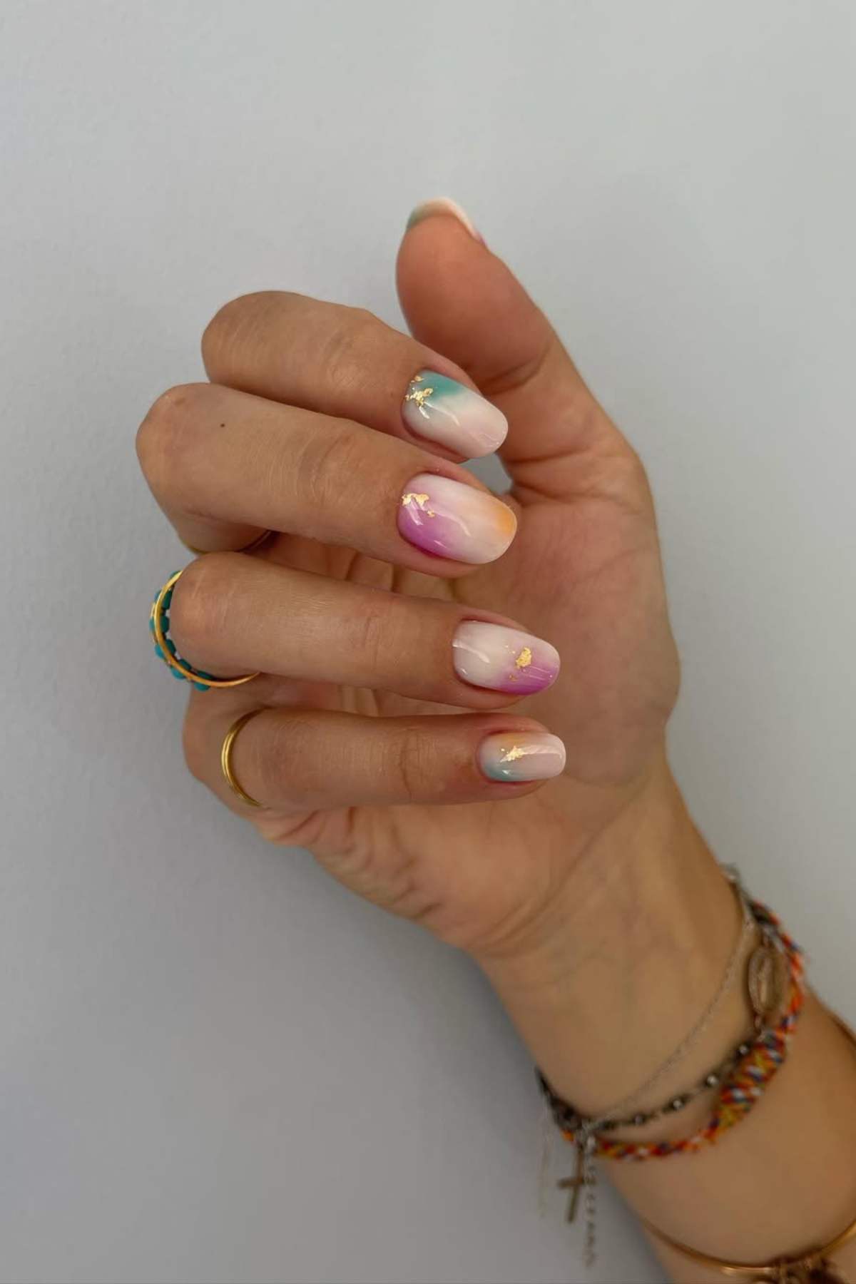 A hand with multicolored pastel nail art and gold accents, wearing several rings and bracelets, shown against a plain light background.