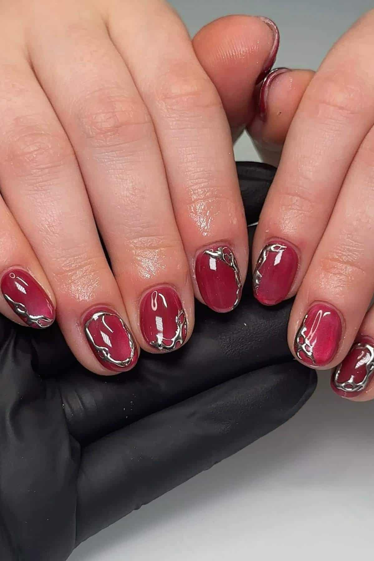 Hands with short, rounded nails painted in deep red polish with silver metallic wavy accents, displayed over a black-gloved hand.