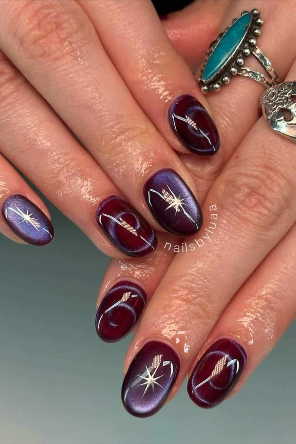 Close-up of hands with almond-shaped nails painted in glossy maroon and purple shades, featuring star and feather designs, accessorized with silver rings.