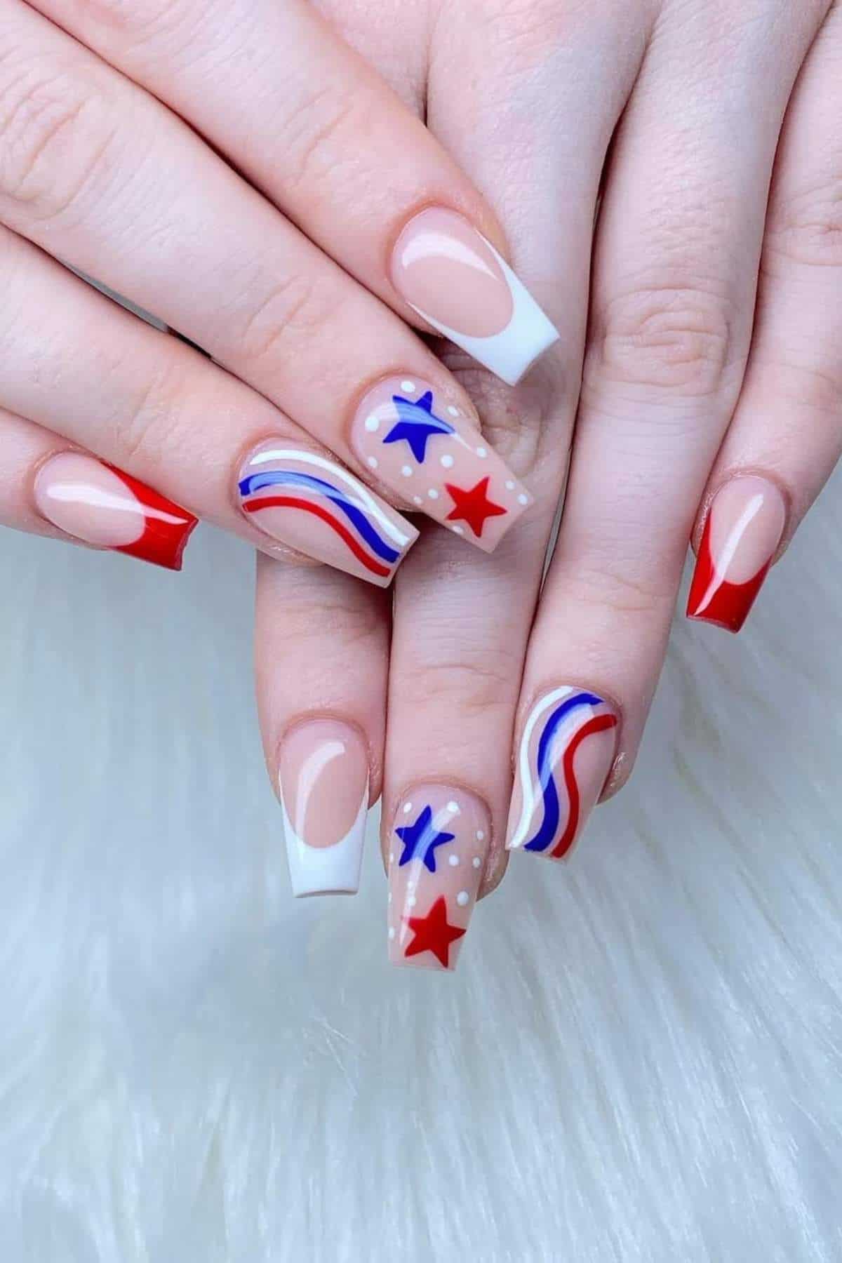 Hands with manicured nails featuring red, white, and blue designs, including stars, stripes, and color blocks, displayed against a white, fluffy background.