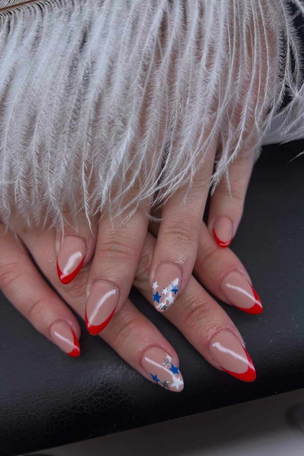 A hand with almond-shaped nails featuring red tips and accent nails decorated with blue and white stars, resting under a white feather.