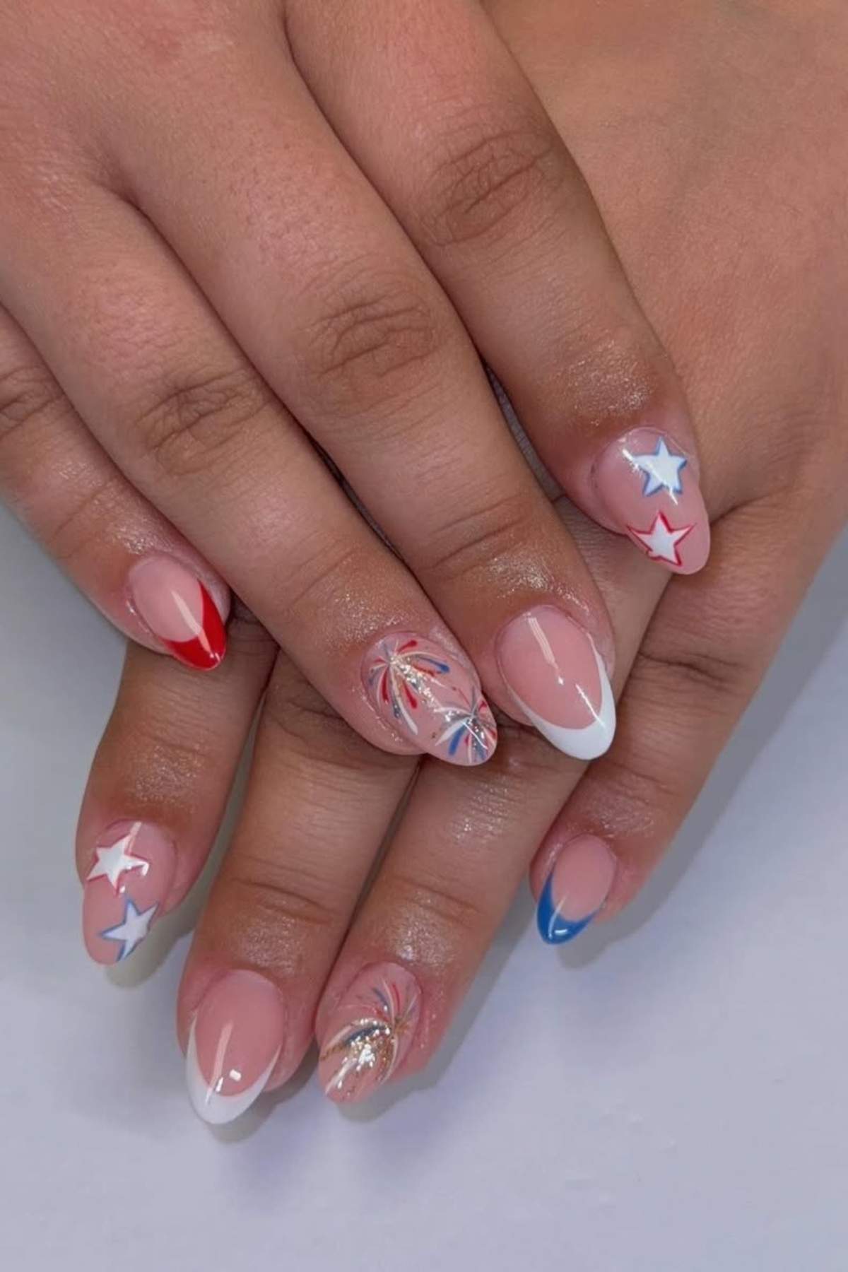 Close-up of hands with manicured nails featuring red, white, and blue designs, stars, and firework patterns on a neutral background.