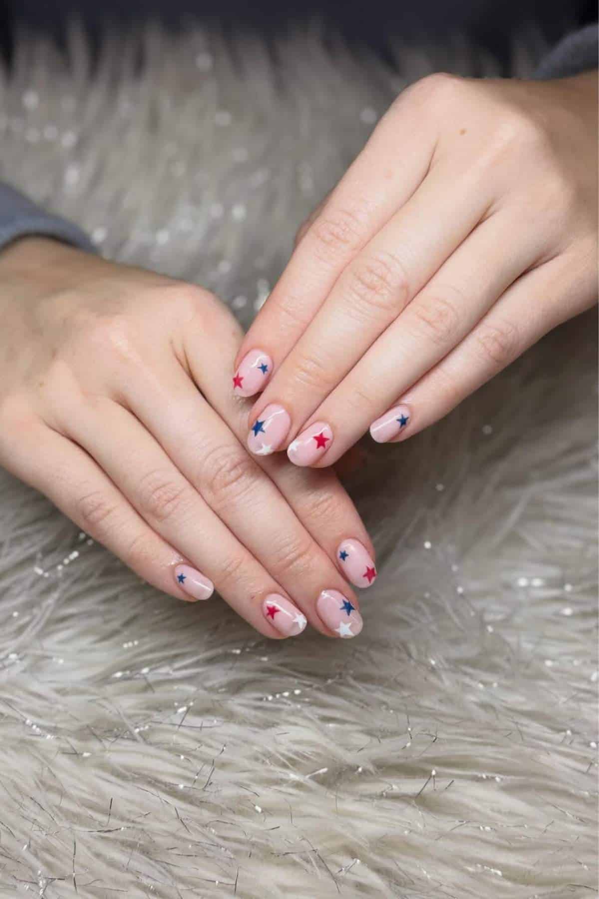 Two hands rest on a textured surface, displaying manicured nails with a nude base and red and blue star decorations on each nail.