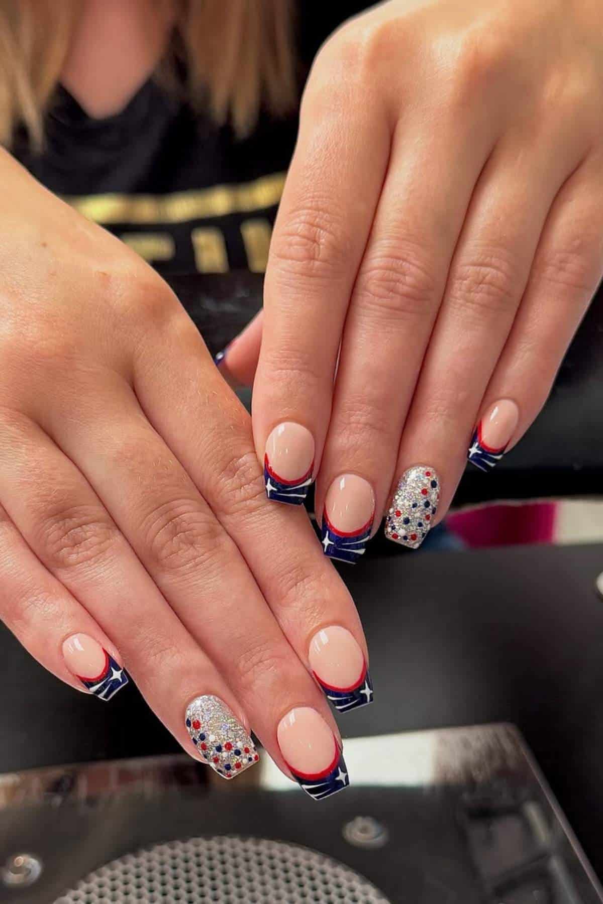 Two hands with manicured nails featuring nude polish, blue and red French tips with white stars, and accent nails with silver glitter and multicolored dots.