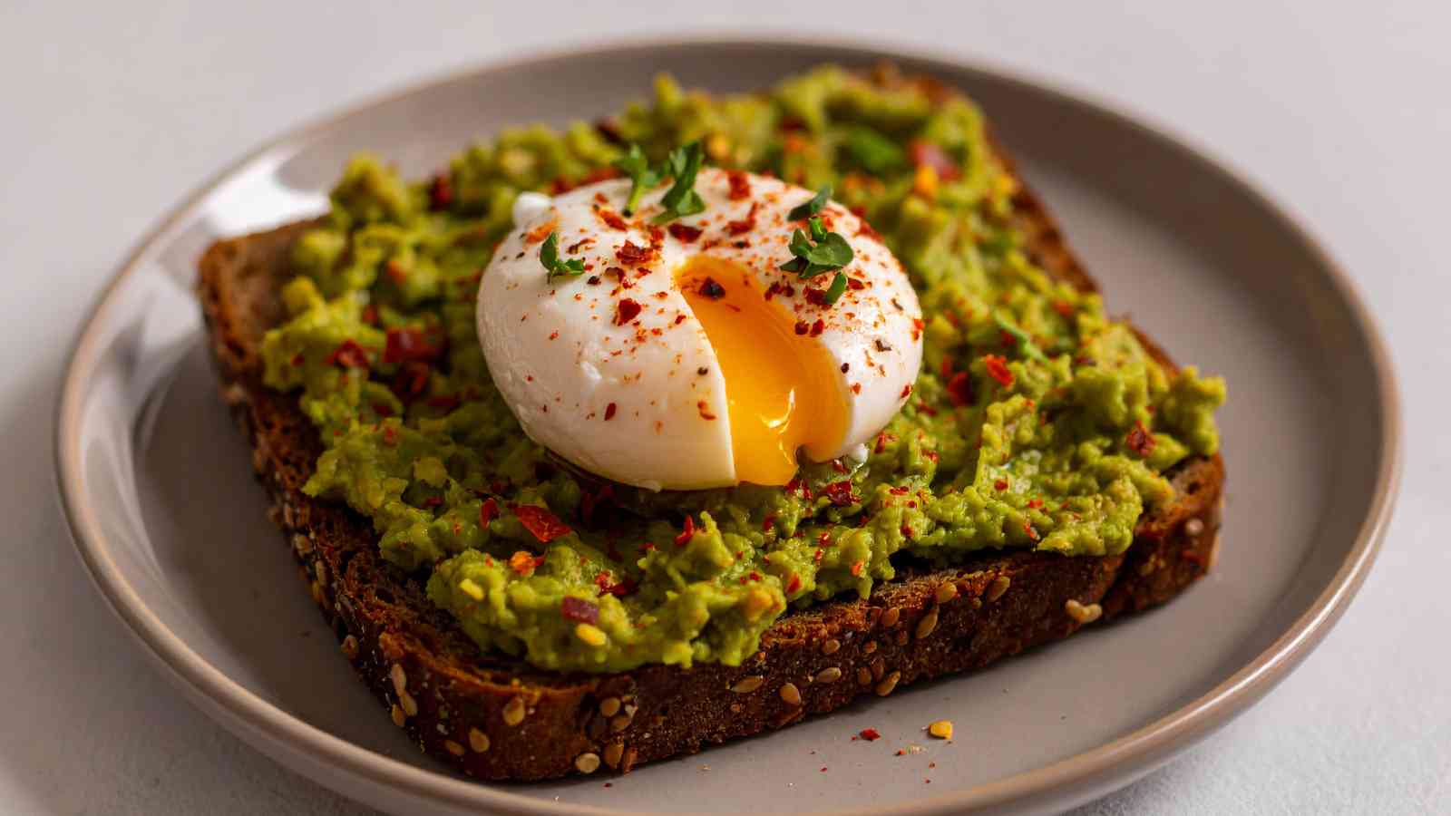 A slice of multigrain toast topped with smashed avocado and a poached egg with a runny yolk, garnished with red pepper flakes and herbs on a beige plate.