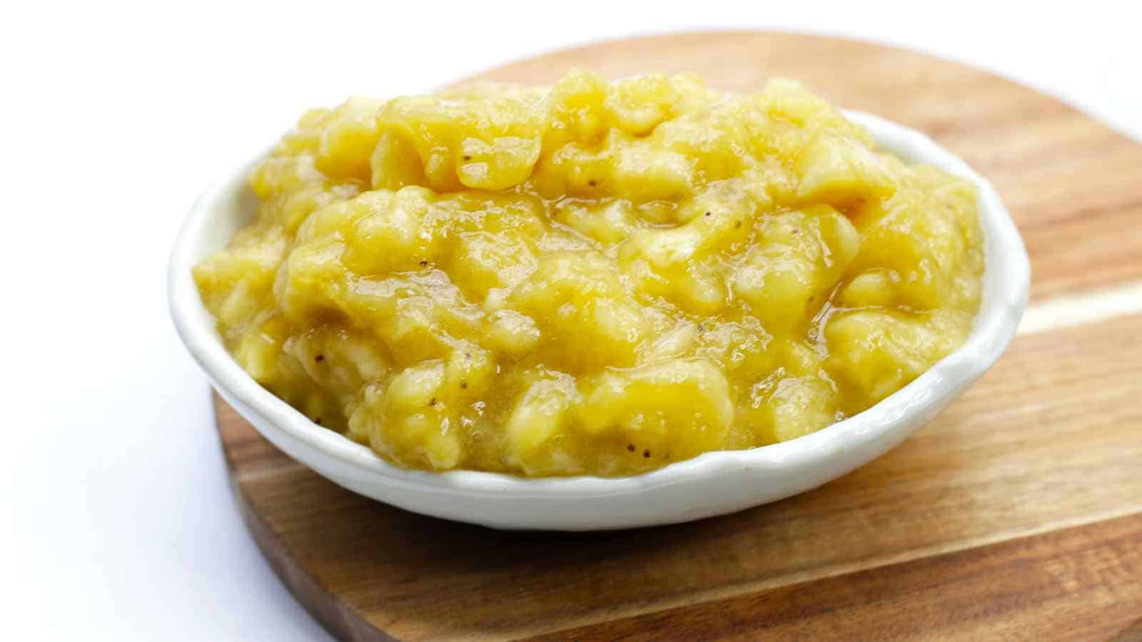 A small white bowl filled with mashed bananas sits on a round wooden board against a white background.