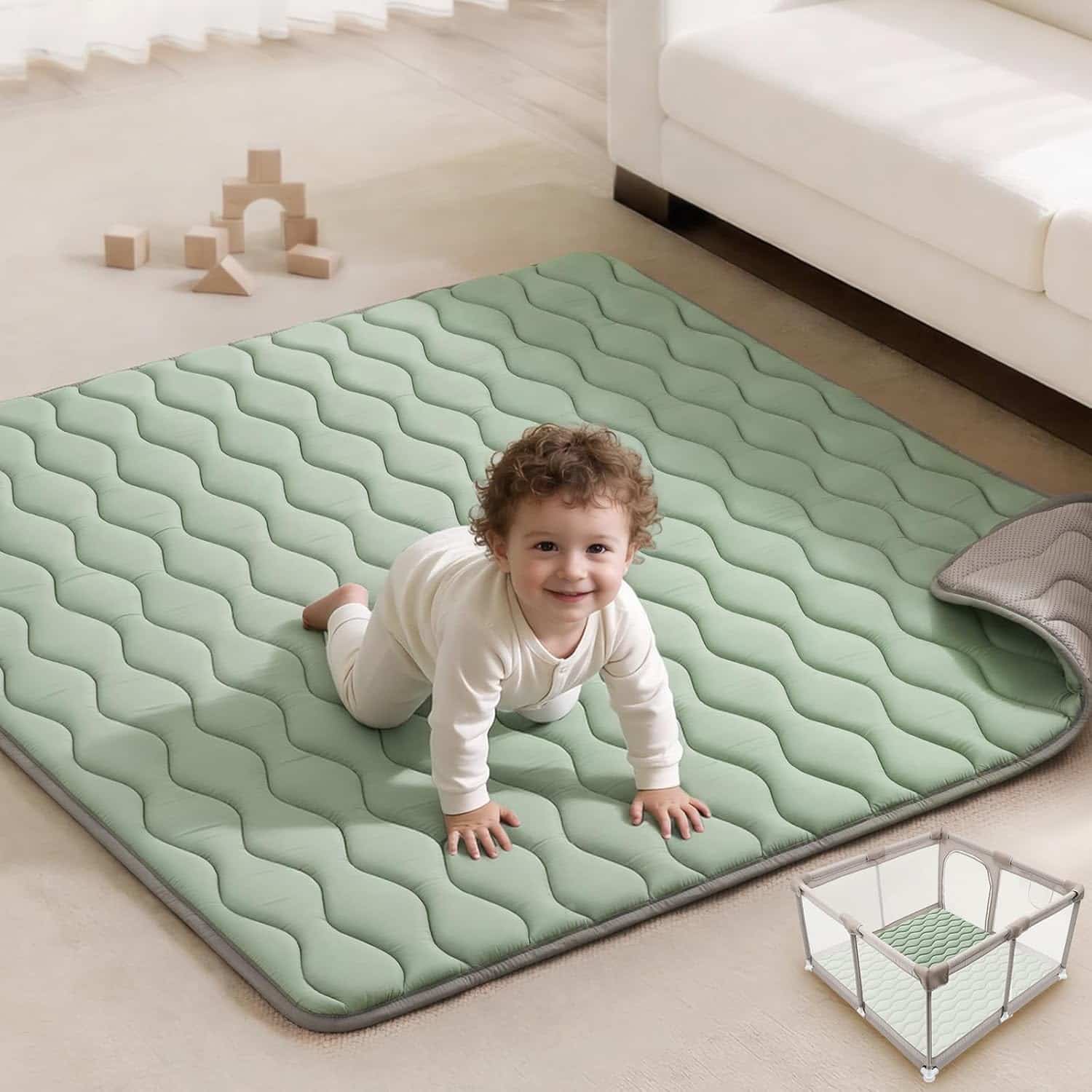 A baby in white clothes crawls on a light green, quilted play mat in a living room, with wooden toys and a playpen visible nearby.