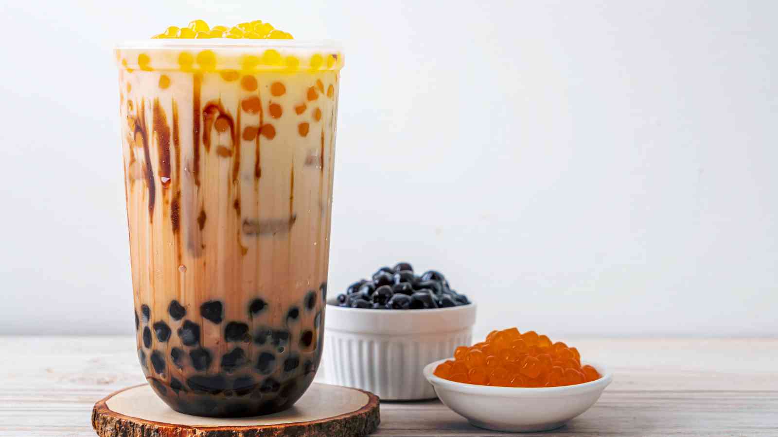 A glass of bubble tea with brown sugar syrup, black tapioca pearls, and yellow popping boba, next to small bowls of black pearls and yellow popping boba on a light surface.
