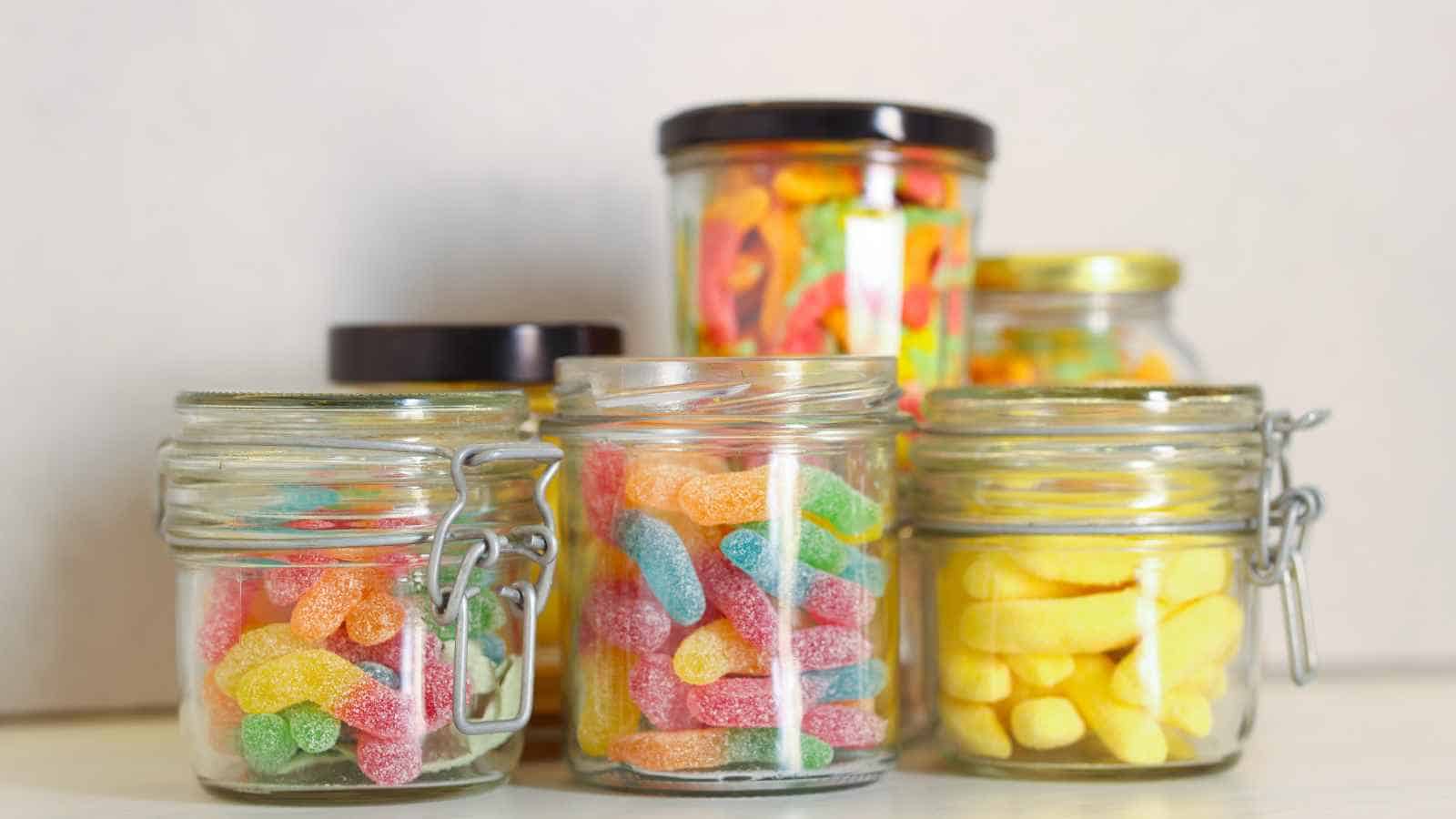 Five glass jars filled with various colorful gummy candies, including rainbow and yellow gummies, are arranged on a light surface against a neutral background.