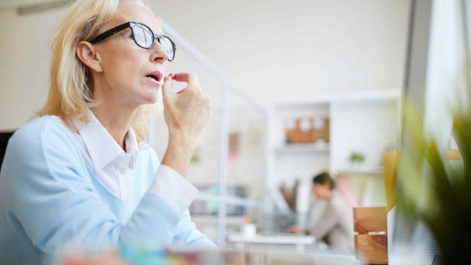 A woman wearing glasses applies lipstick while sitting at a desk in an office, with another person working in the background.