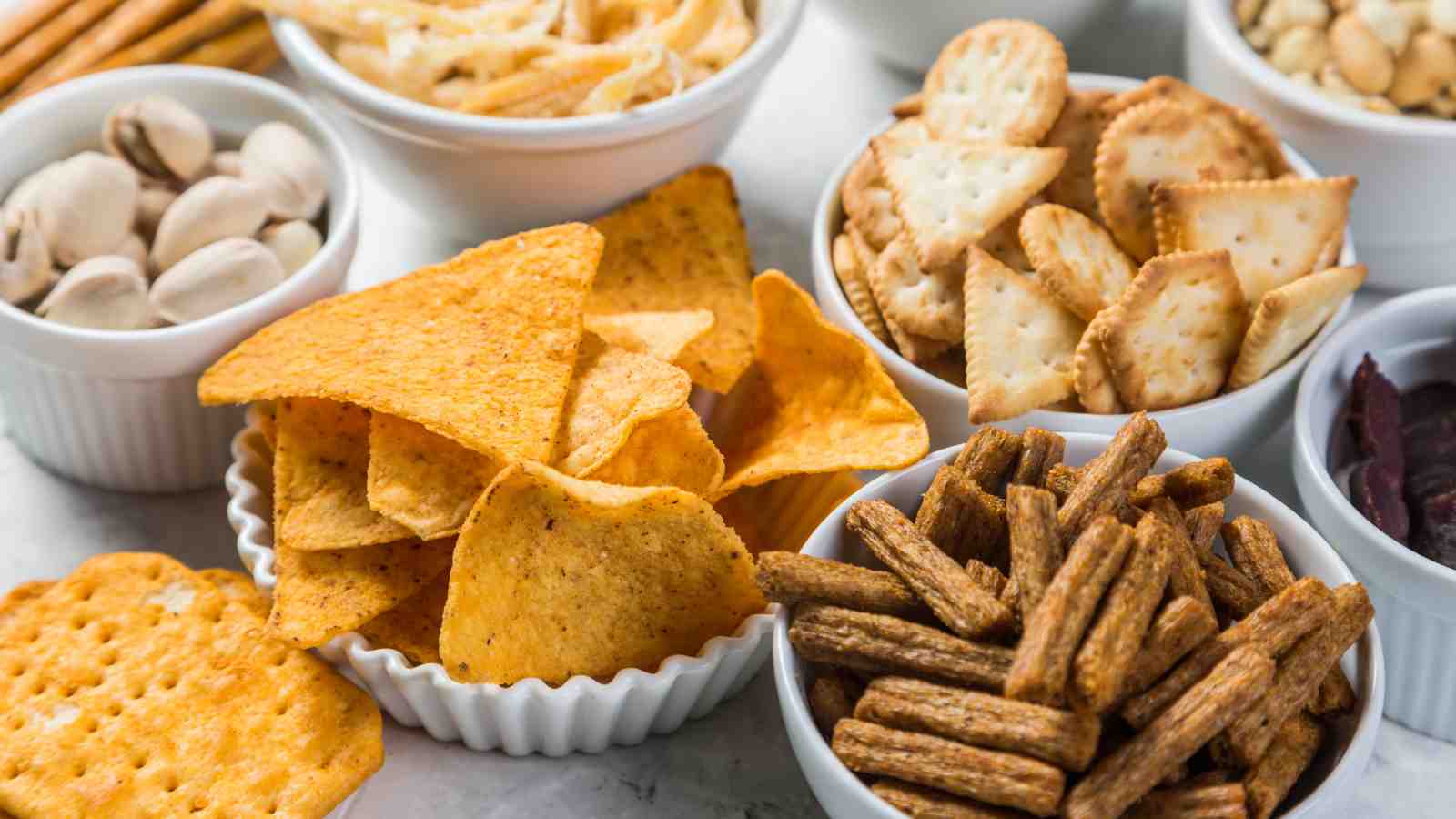 An assortment of snacks including tortilla chips, crackers, breadsticks, pistachios, and other finger foods arranged in bowls on a table.