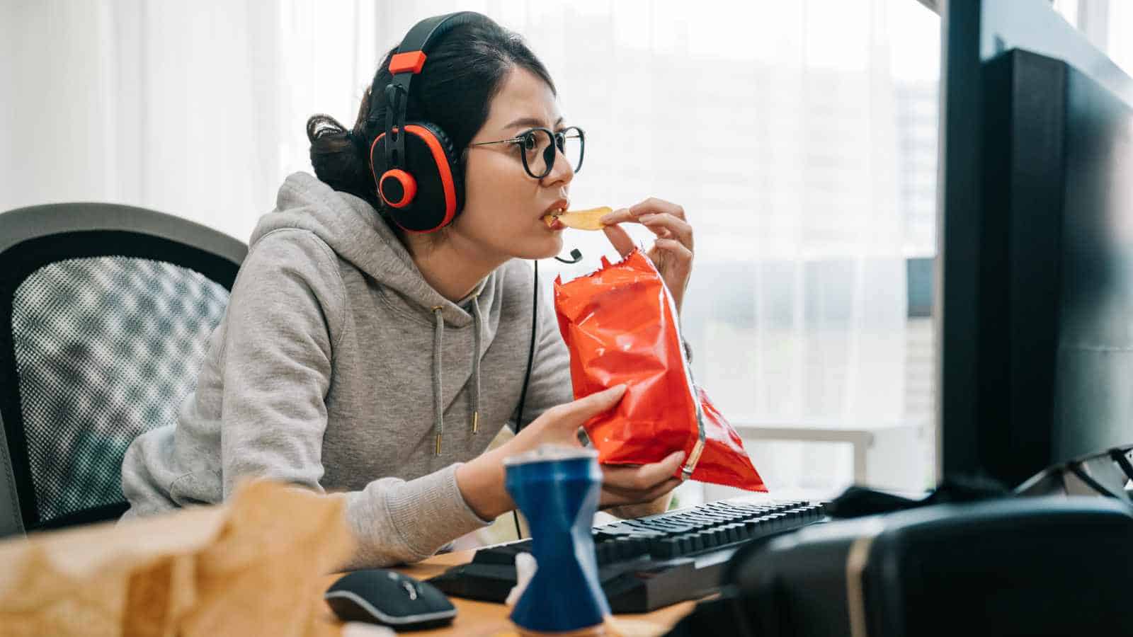 A person wearing headphones and glasses eats chips while using a computer at a desk cluttered with a keyboard, mouse, and a drink.