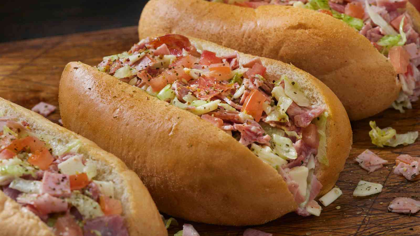 Three hoagie sandwiches filled with chopped deli meats, lettuce, tomato, cheese, and seasonings on a wooden surface.