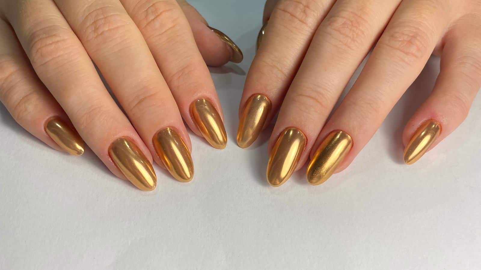 Two hands with almond-shaped nails painted in shiny metallic gold polish, shown against a plain white background.