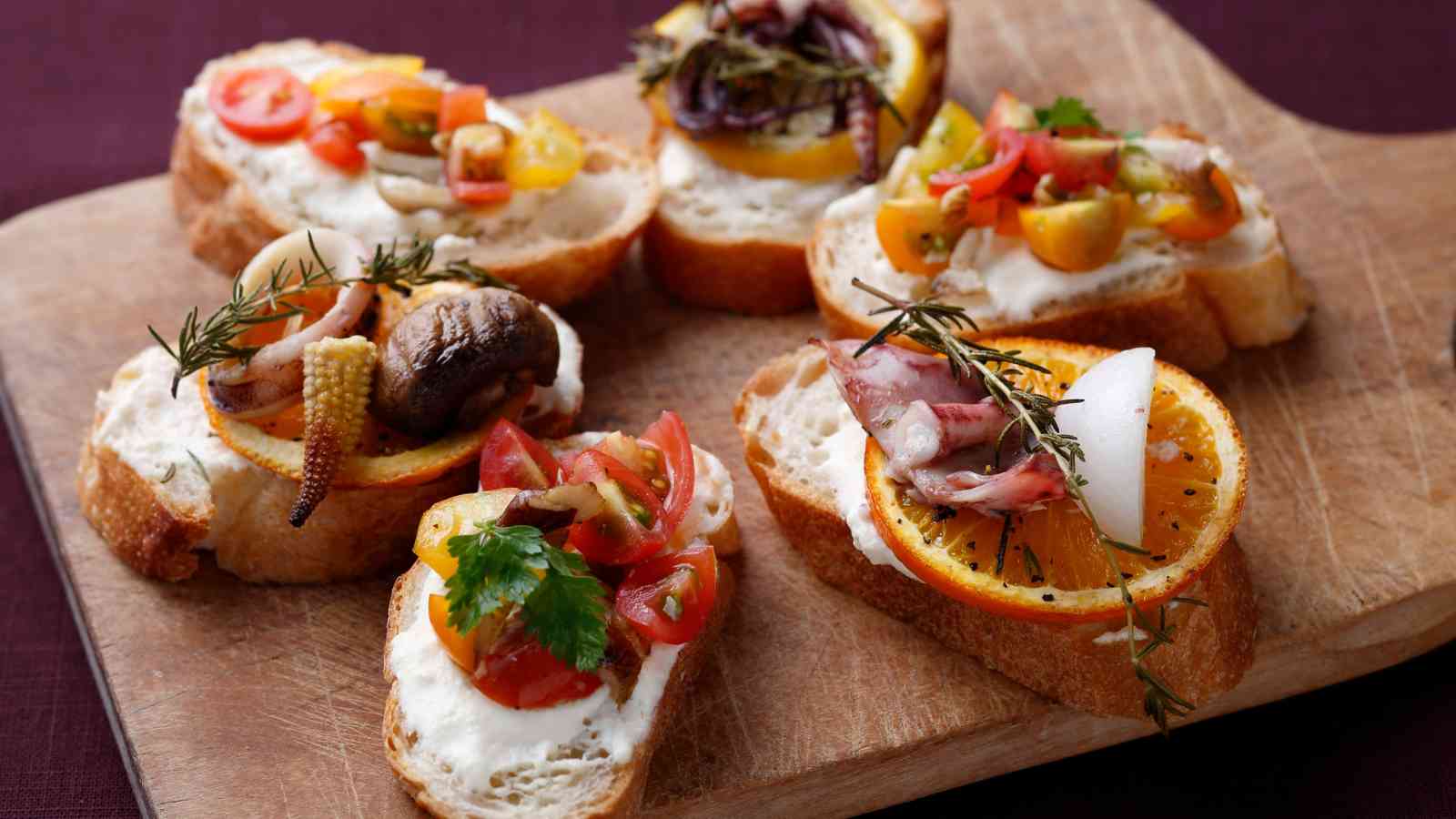 Five slices of bread topped with various vegetables, herbs, and seafood are arranged on a wooden cutting board.