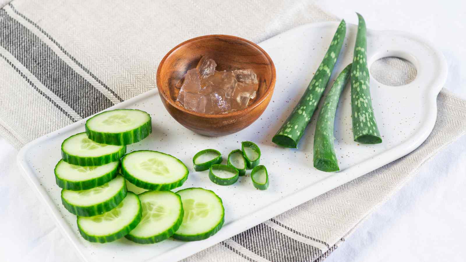 Sliced cucumber, aloe vera leaves, and a wooden bowl of aloe gel arranged on a white tray atop a striped cloth.