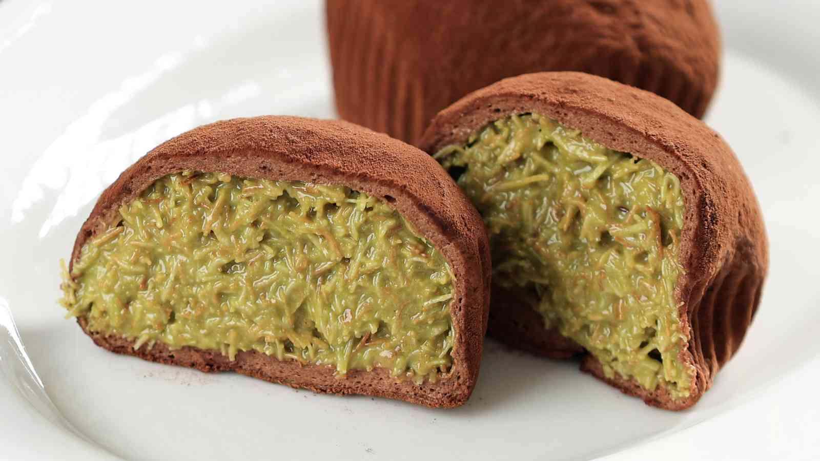 A chocolate-coated dessert cut in half reveals a green, textured filling, served on a white plate.