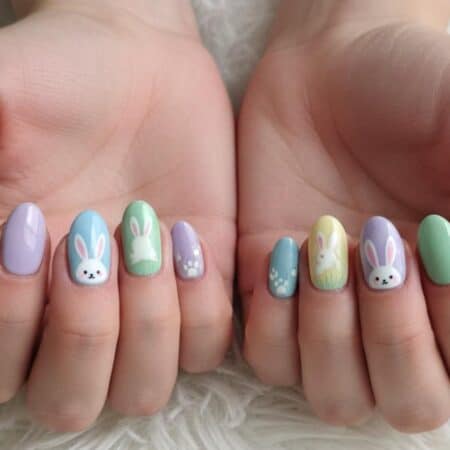 Hands with pastel-colored nail art featuring painted bunny faces, bunny ears, and paw prints on a white textured surface.