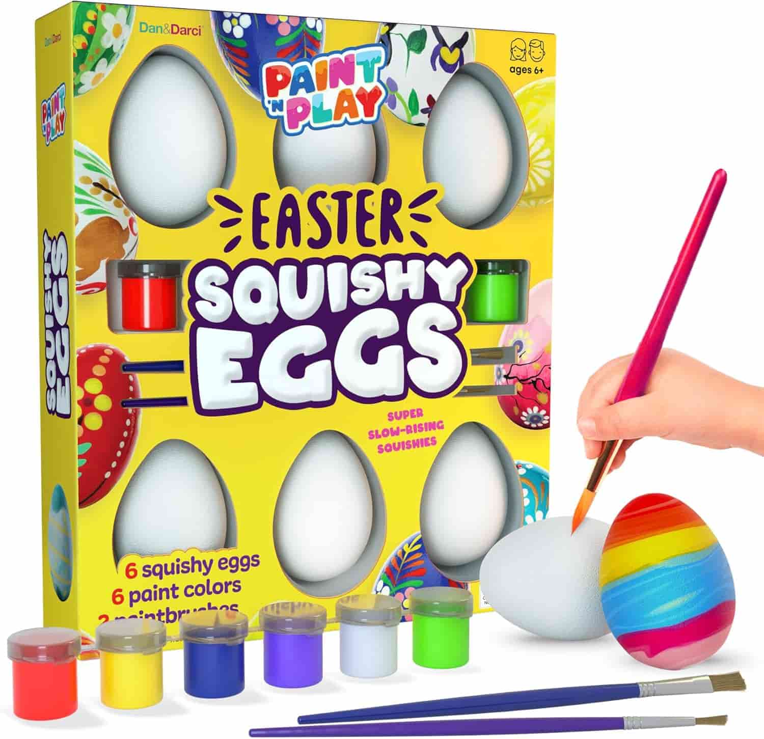 Paint kit with six white squishy eggs, six paint colors, three brushes, and a hand painting a rainbow design on one egg; packaging reads “Easter Squishy Eggs.”.