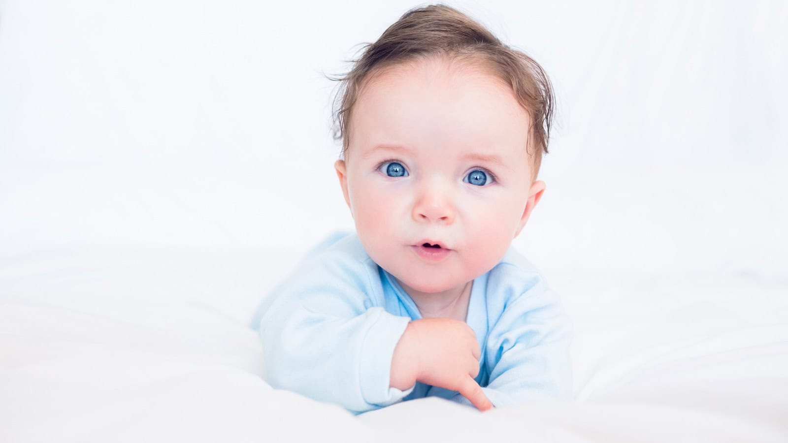A baby with blue eyes and light brown hair lies on a white bed, wearing a light blue outfit and looking directly at the camera.