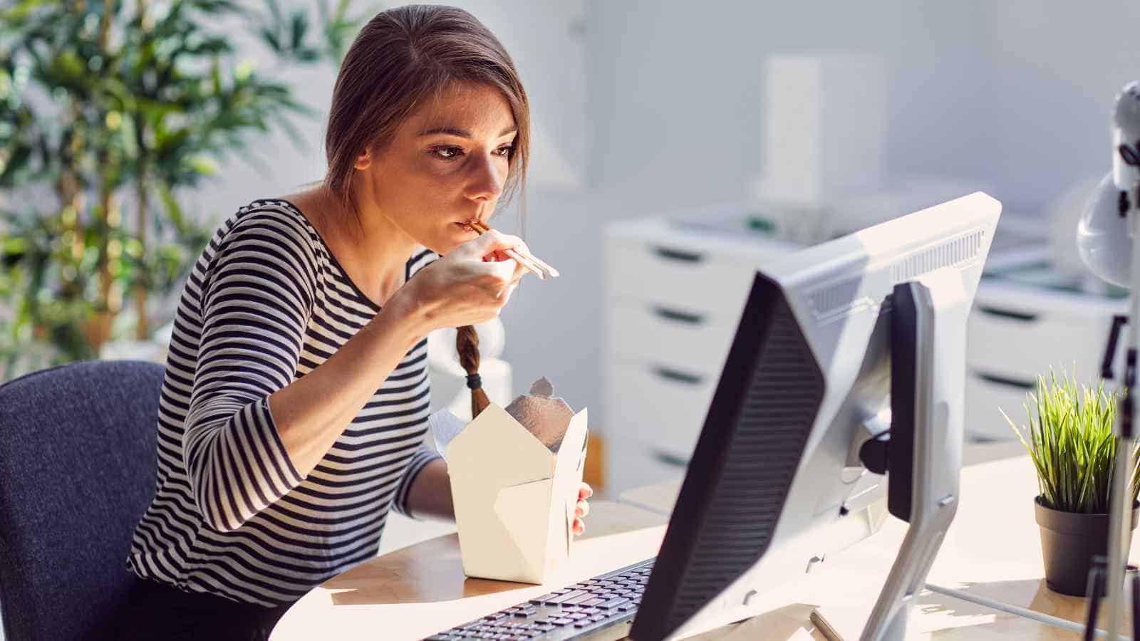 A person in a striped shirt sits at a desk, eating takeout food with chopsticks while looking at a computer monitor in a bright, plant-filled office.