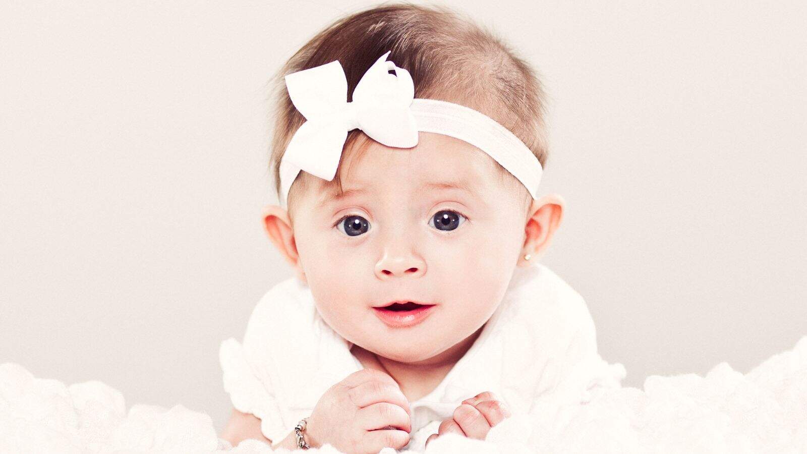 A baby wearing a white headband with a bow and a white outfit lies on a soft white surface, looking towards the camera.
