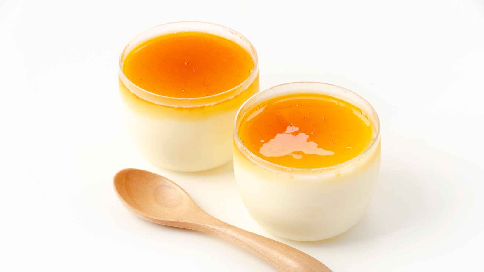 Two glass cups of caramel pudding are placed on a white surface next to a wooden spoon.