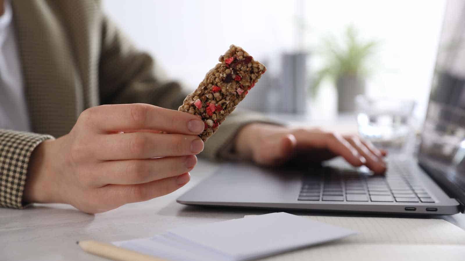 Person holding a granola bar in one hand while using a laptop with the other, with a notepad and pen on the table.