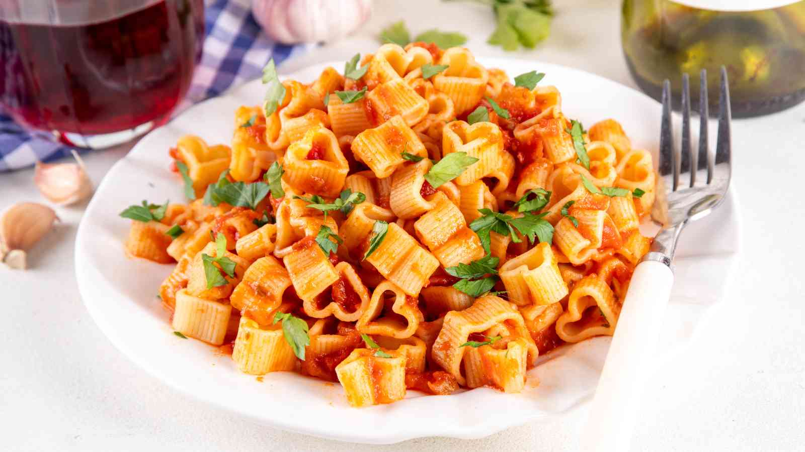 A white plate of pasta with tomato sauce and parsley, served with a fork. A glass of red wine, garlic, and a bottle are visible in the background.