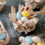 A close-up of a homemade edible bird nest treat made from cereal sticks and white coating, filled with candy eggs, on a textured surface.