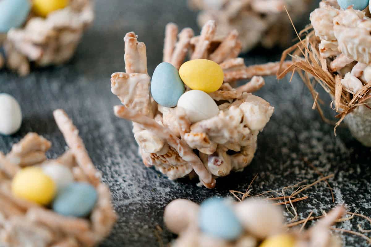 A close-up of a homemade edible bird nest treat made from cereal sticks and white coating, filled with candy eggs, on a textured surface.