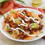A plate of nachos topped with ground beef, melted cheese, pickle slices, sour cream, and chopped green onions, with tortilla chips and green onions beside the plate.