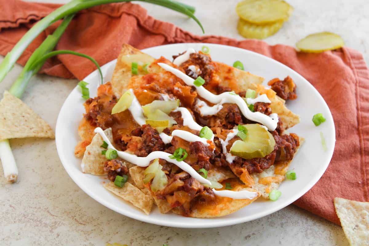 A plate of nachos topped with ground beef, melted cheese, pickle slices, sour cream, and chopped green onions, with tortilla chips and green onions beside the plate.