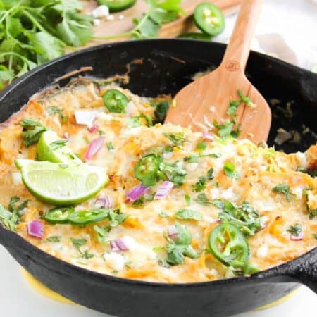 A cast iron skillet filled with a cheesy, baked enchilada topped with lime wedges, sliced jalapeños, chopped red onion, and fresh cilantro. A wooden spoon rests in the skillet.