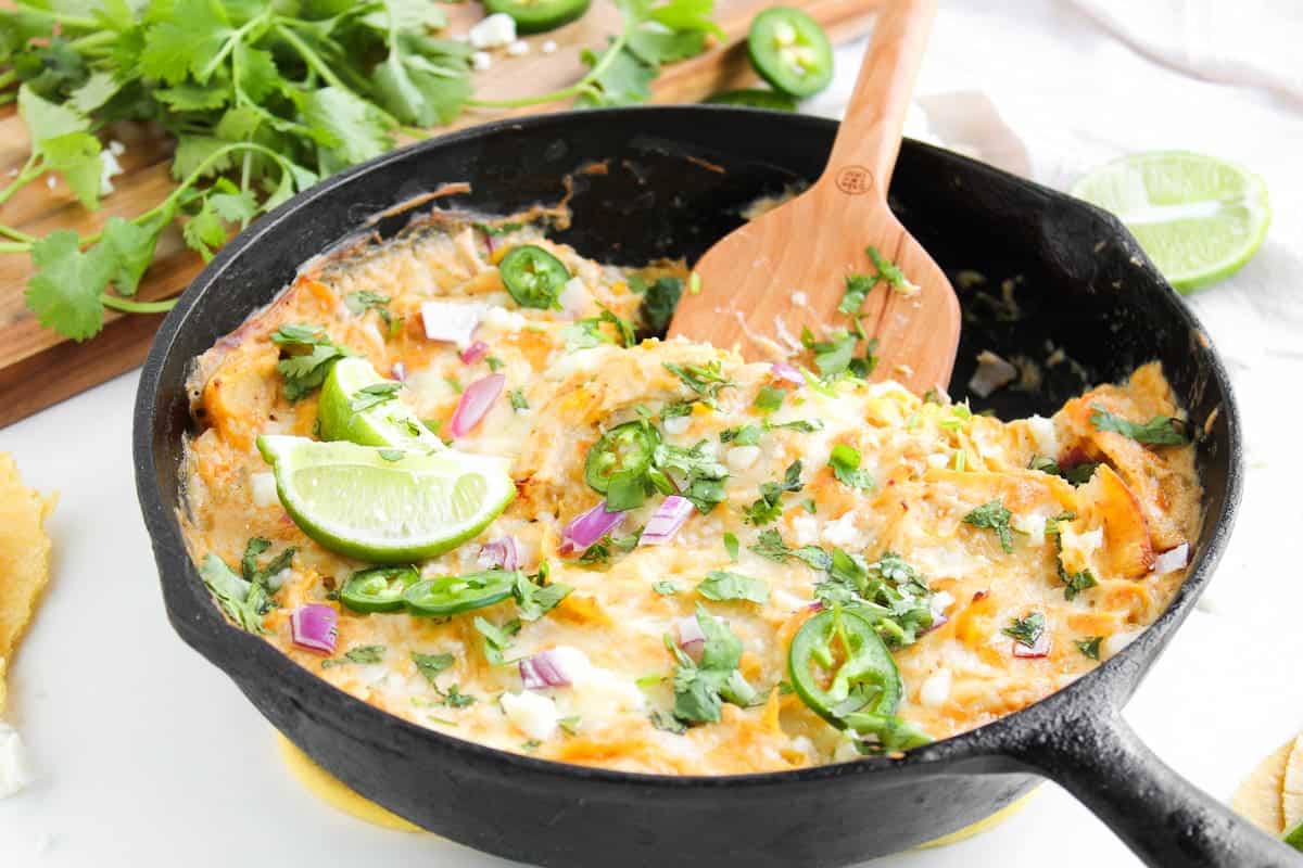 A cast iron skillet filled with a cheesy, baked enchilada topped with lime wedges, sliced jalapeños, chopped red onion, and fresh cilantro. A wooden spoon rests in the skillet.