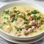 A bowl of creamy soup with sausage, kale, potatoes, and bacon in a light broth, served in a white bowl on a cloth napkin.
