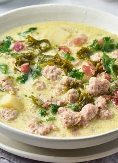 A bowl of creamy soup with sausage, kale, potatoes, and bacon in a light broth, served in a white bowl on a cloth napkin.