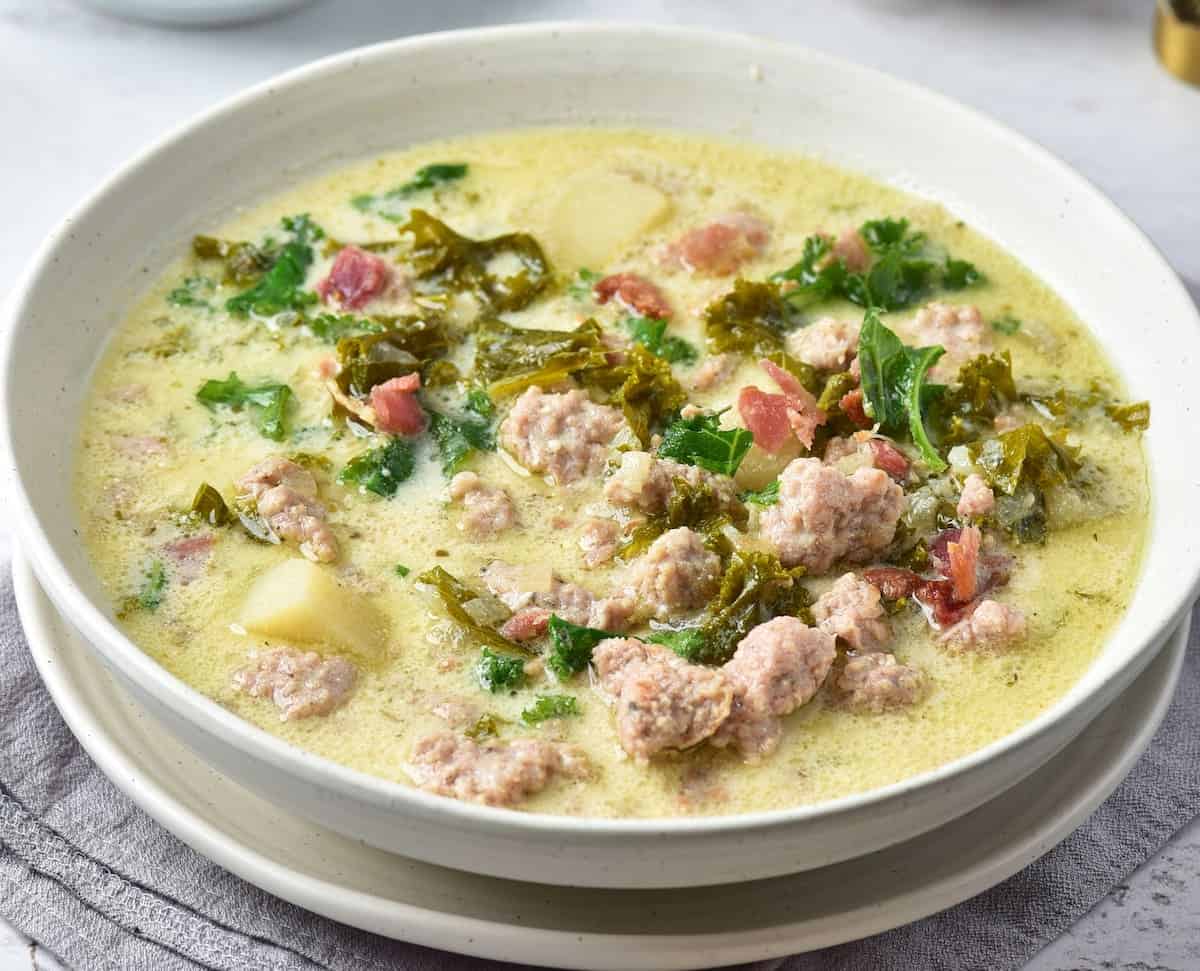 A bowl of creamy soup with sausage, kale, potatoes, and bacon in a light broth, served in a white bowl on a cloth napkin.