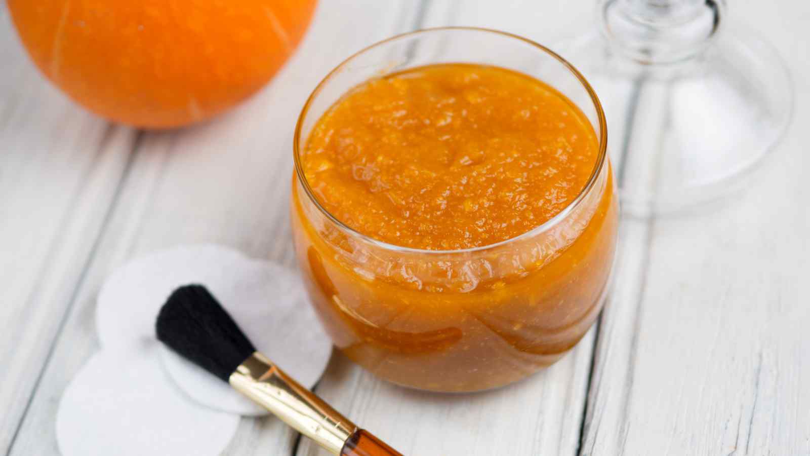 A glass jar filled with orange-colored gel or jam is on a white wooden surface next to a makeup brush, cotton pads, and a partial orange fruit.
