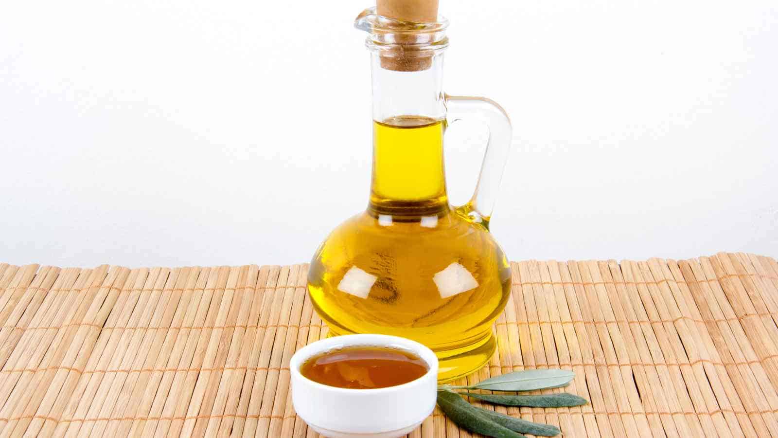 A glass bottle of olive oil with a cork, a small white bowl containing oil, and a few olive leaves, all placed on a bamboo mat.